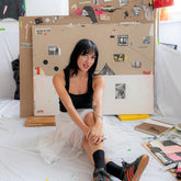 Artist Poppy Faun sits on the floor of her studio with her legs crossed. A selection of her paintings are resting behind her.